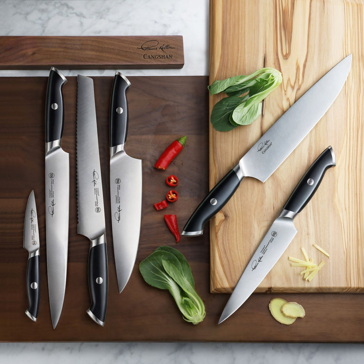 Seven black-handled knives with stainless steel blades arranged on wooden cutting boards with sliced chili, bok choy, and gin
