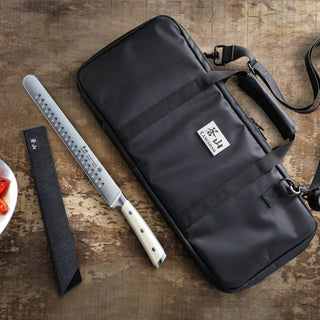 
                    
                      Black nylon knife bag with strap and handles, lying flat on a wooden surface next to a long knife with a white handle and bla
                    
                  