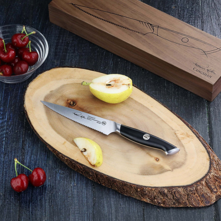 Serrated utility knife with black handle on wooden cutting board, next to sliced pear and bowl of cherries.