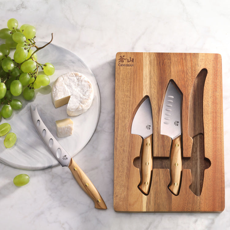 Three olive wood-handled cheese knives on an acacia wood board next to a marble plate with cheese and green grapes.