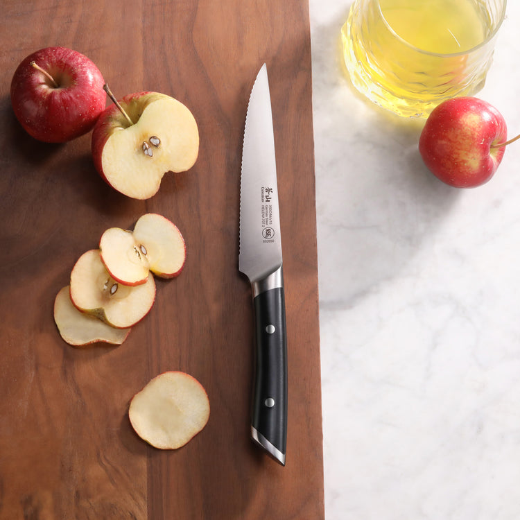 Serrated utility knife with black handle on wooden cutting board next to sliced and whole apples and a glass of apple juice.