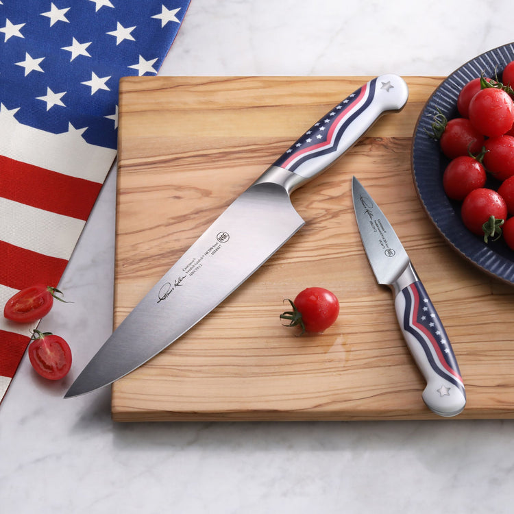 Two knives with American flag design handles on a wooden cutting board, surrounded by cherry tomatoes and a flag cloth.
