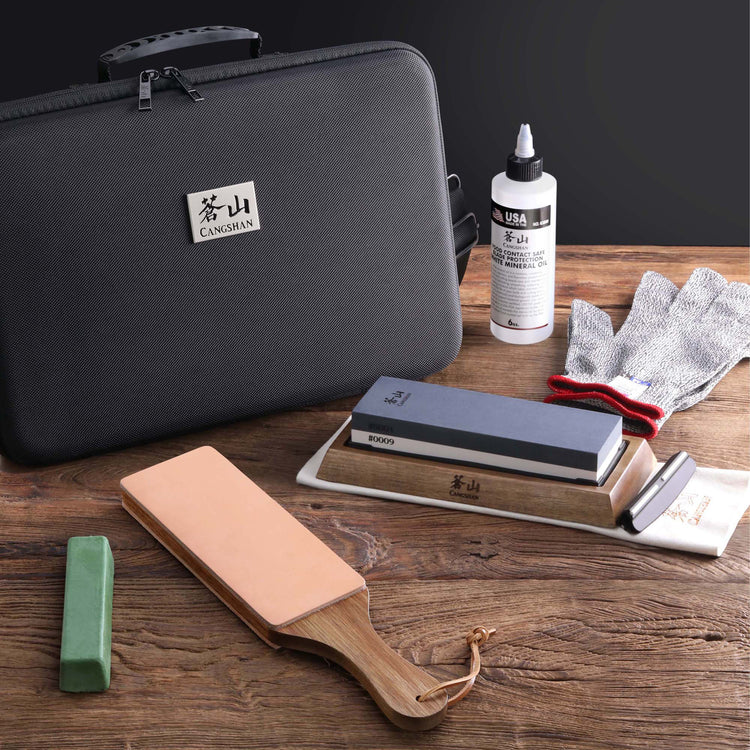 Knife sharpening stones, honing oil, protective glove, leather strop, and carrying case on wooden surface.
