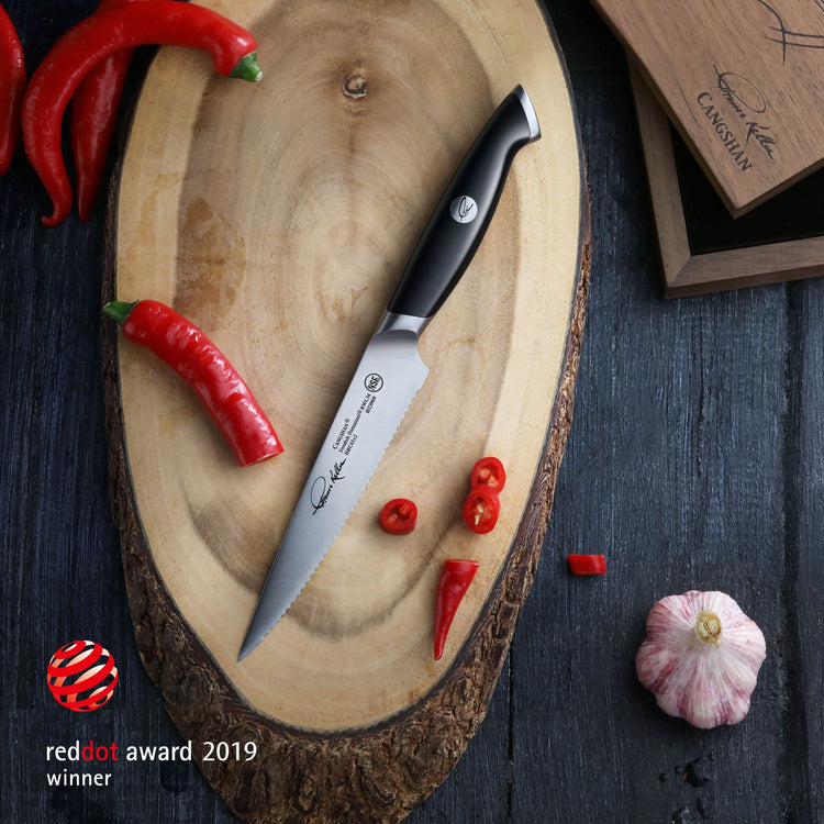5-inch serrated utility knife with black handle on wooden board, surrounded by red chili peppers and garlic bulb.