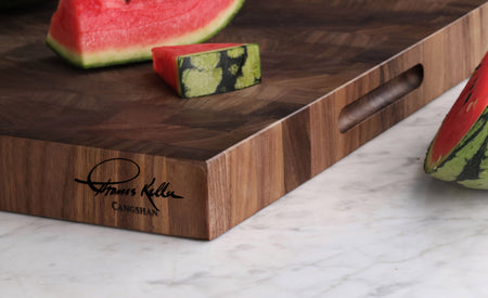 Walnut end-grain cutting board with engraved Thomas Keller signature and watermelon slices on top and beside it.