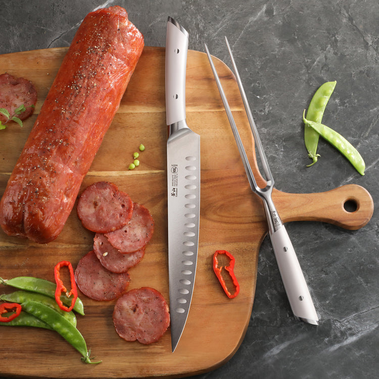 Carving knife and fork with white handles on wooden board next to sliced sausage and vegetables.