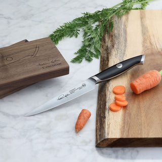 
                    
                      Black-handled fine-edge utility knife with engraved blade, placed on a wooden cutting board next to sliced carrots and a wood
                    
                  