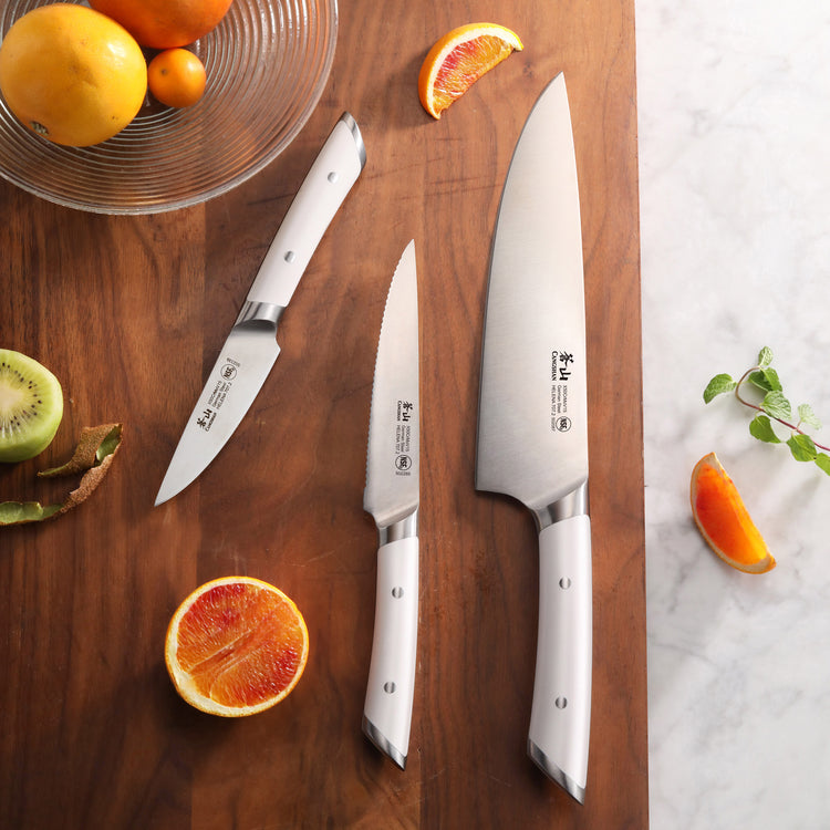 Three stainless steel kitchen knives with white handles arranged on a wooden cutting board next to sliced citrus fruits and a