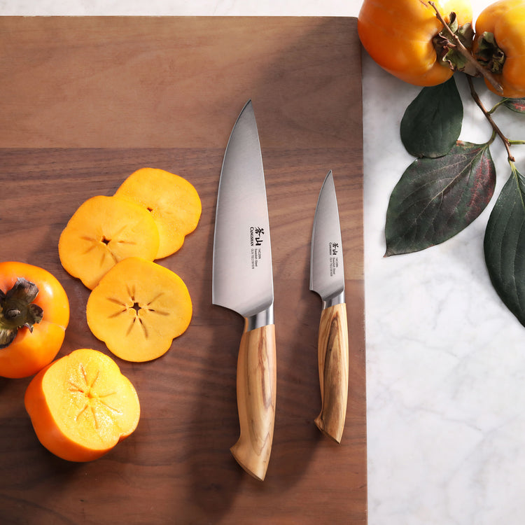 Two kitchen knives with wooden handles on a cutting board next to sliced and whole persimmons.