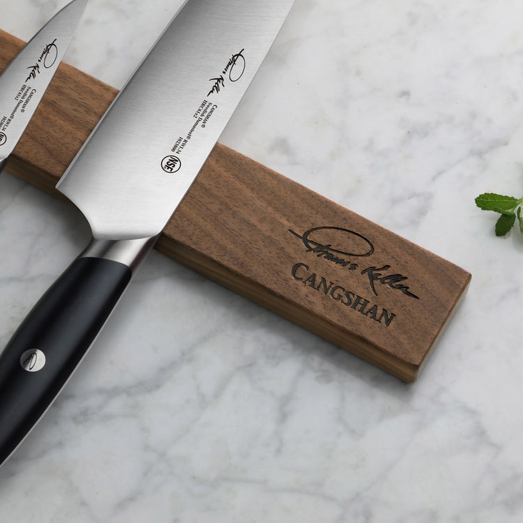 Two stainless steel knives with black handles resting on a wooden magnetic knife holder with engraved signature and brand nam