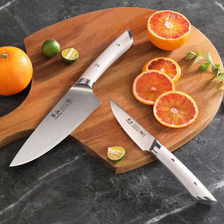 Two stainless steel knives with white handles on a wooden board, alongside sliced and whole citrus fruits.