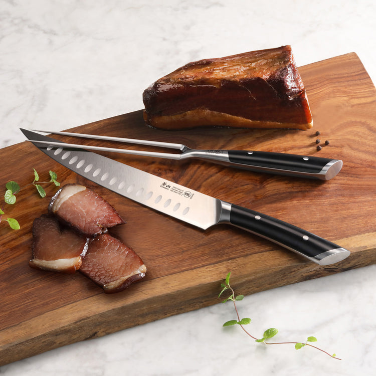 Two-piece carving set with a slicing knife and fork on a wooden board next to sliced and whole cooked meat.