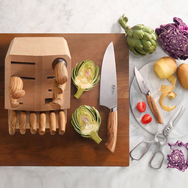 Wooden knife block with knives, a cutting board, halved artichokes, peeled potatoes, cherry tomatoes, scissors, and purple ca