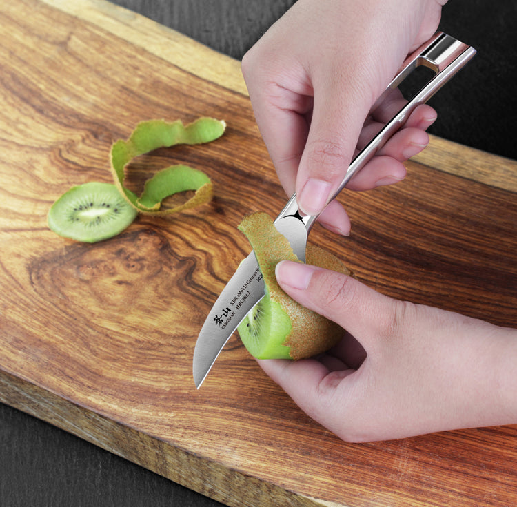 Hands peeling a kiwi fruit with a small curved peeling knife on a wooden cutting board.