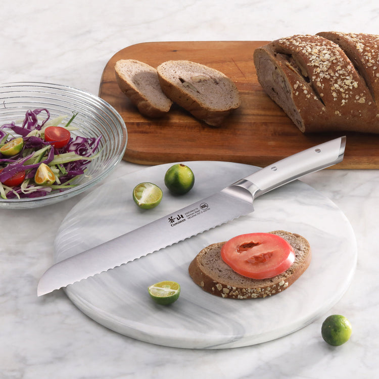 Serrated bread knife with stainless steel handle on a marble board, next to sliced bread, lime halves, and a salad bowl.