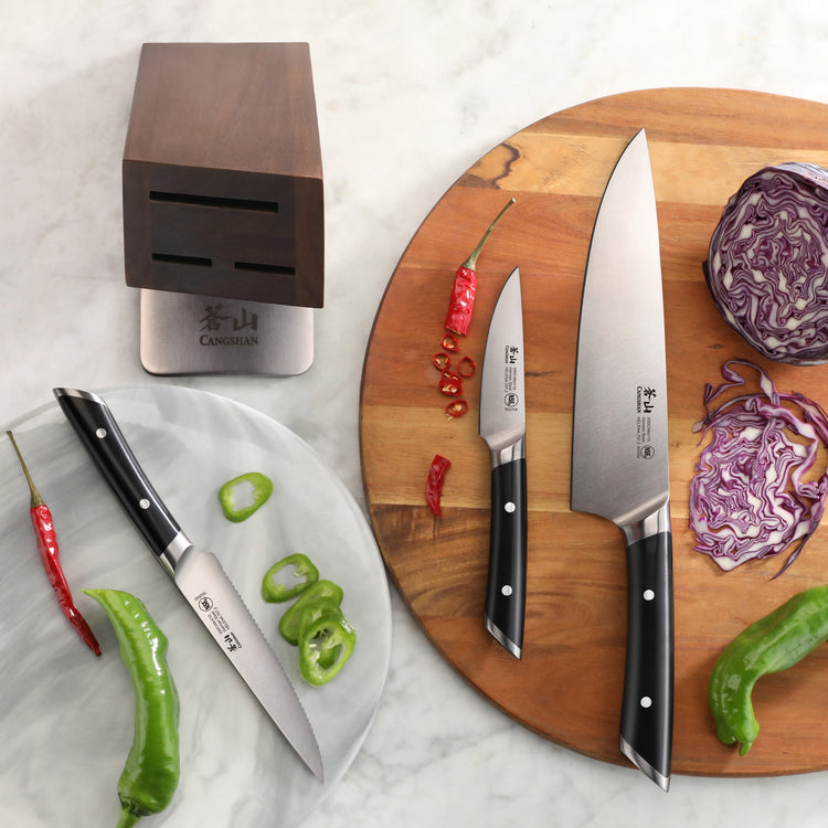 Four knives with black handles displayed with sliced peppers and cabbage next to a wooden knife block on a marble surface.