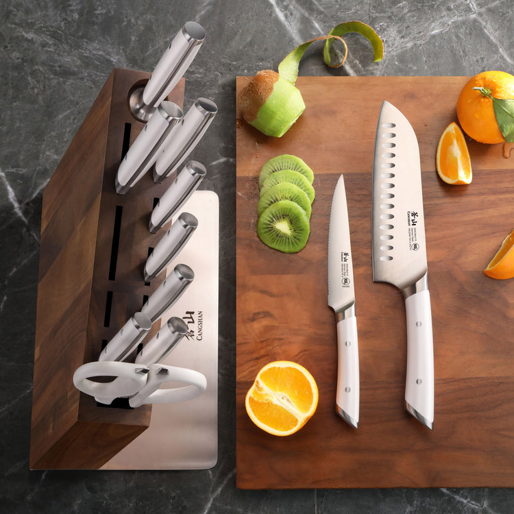 Knife block set with six knives and scissors on a wooden block, two knives on a cutting board with sliced kiwi and orange.