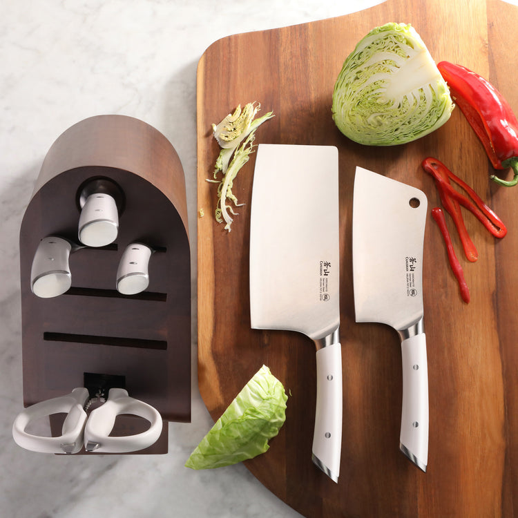 Two forged German steel cleaver knives with white handles on a wooden cutting board next to an acacia wood knife block and ve