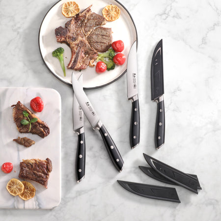 Four black-handled steak knives with sheaths next to cooked steak and vegetables on white plates on a marble surface.