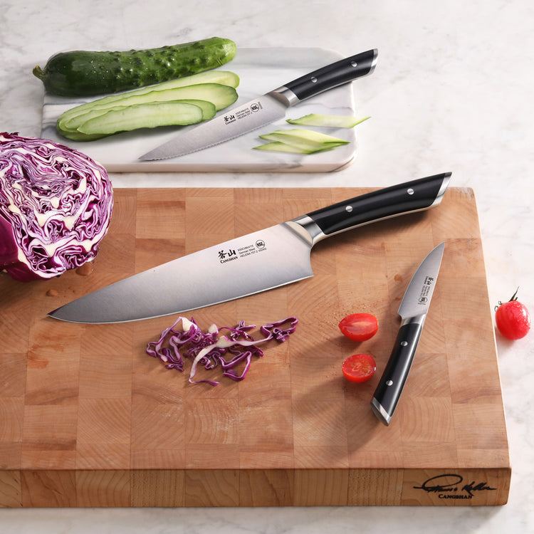 Two kitchen knives with black handles on wooden cutting boards, one slicing cucumber and the other chopping red cabbage and c