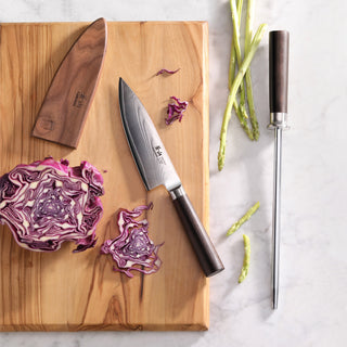 
                    
                      6-inch chef's knife with wooden handle and sheath on cutting board with sliced red cabbage and sharpening steel nearby.
                    
                  