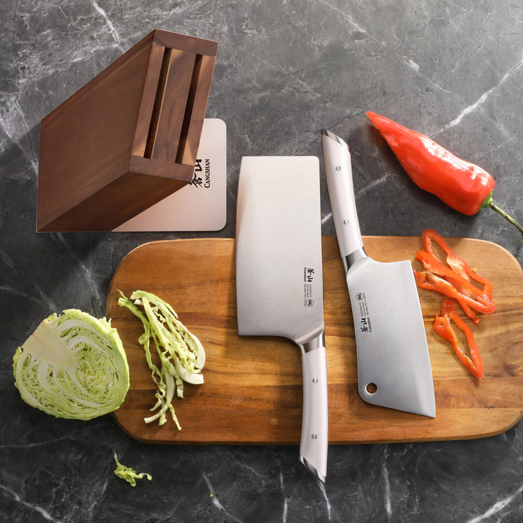 Two cleaver knives with white handles on a wooden cutting board, next to sliced cabbage, red pepper, and an empty acacia wood