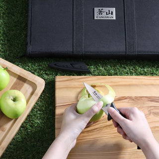 
                    
                      Hands peeling a green apple with a small knife on a wooden cutting board, next to a black knife bag on grass.
                    
                  