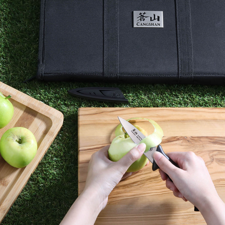 Hands peeling a green apple with a small knife on a wooden cutting board, next to a black knife bag on grass.
