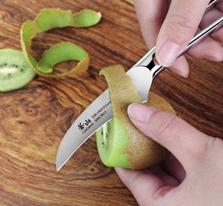 
                    
                      Hand peeling a kiwi with a small curved peeling knife on a wooden surface.
                    
                  
