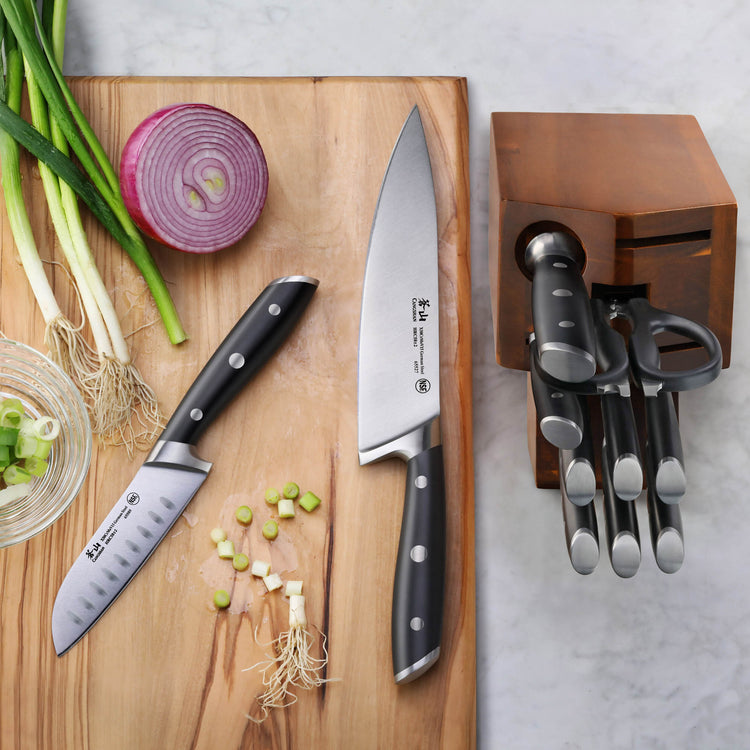 Two knives with black handles on a wooden cutting board next to chopped green onions and a halved red onion, with a wooden kn