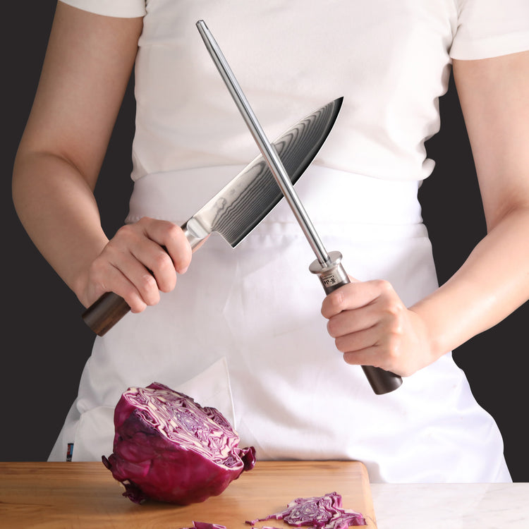 Person holding a knife and a honing steel with a halved red cabbage on a cutting board.