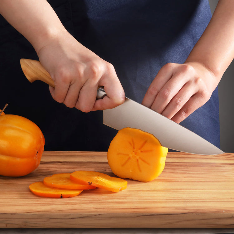 Hands slicing a ripe persimmon on a wooden cutting board with a chef's knife featuring a wooden handle.