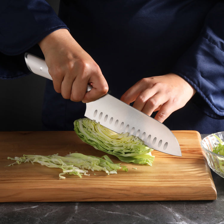 Hand holding a 7-inch Santoku knife slicing a wedge of cabbage on a wooden cutting board.