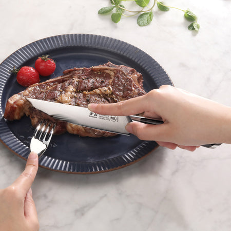 Hand holding a steak knife cutting a cooked steak on a blue plate with two cherry tomatoes and a fork holding the steak.