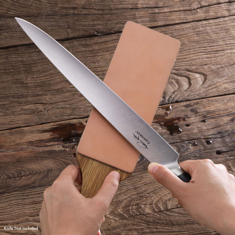 Hands holding a sharpening stone and a large kitchen knife blade on a wooden surface.