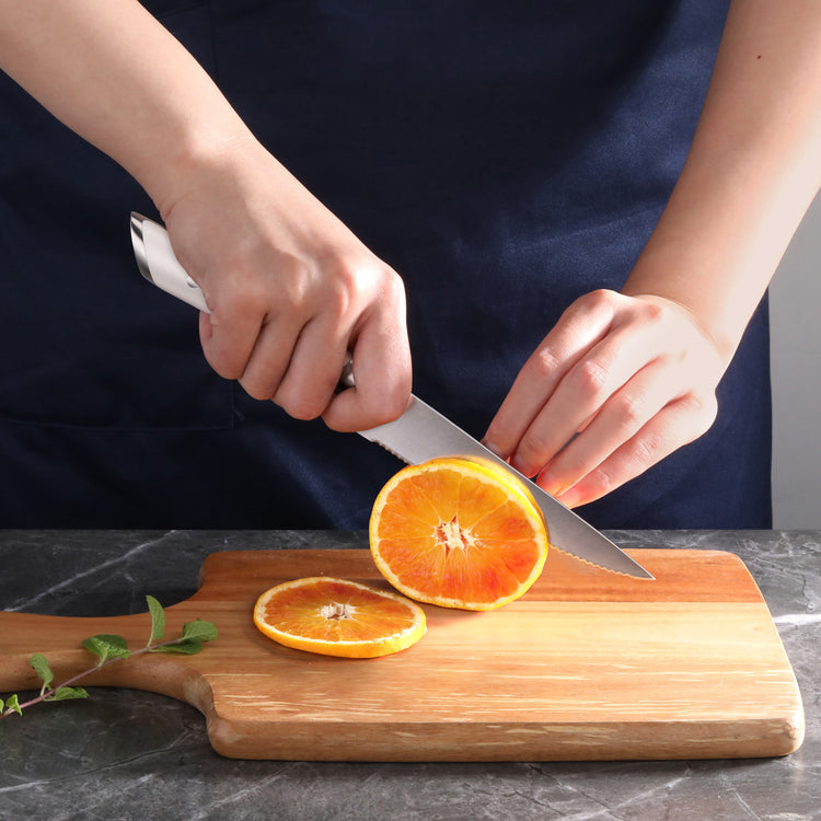 Hands slicing an orange on a wooden cutting board with a serrated utility knife.