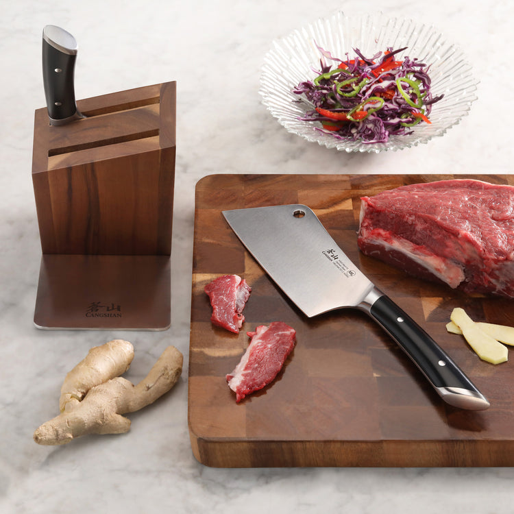 Cleaver knife in acacia wood block next to cutting board with raw meat, ginger, and a bowl of salad.