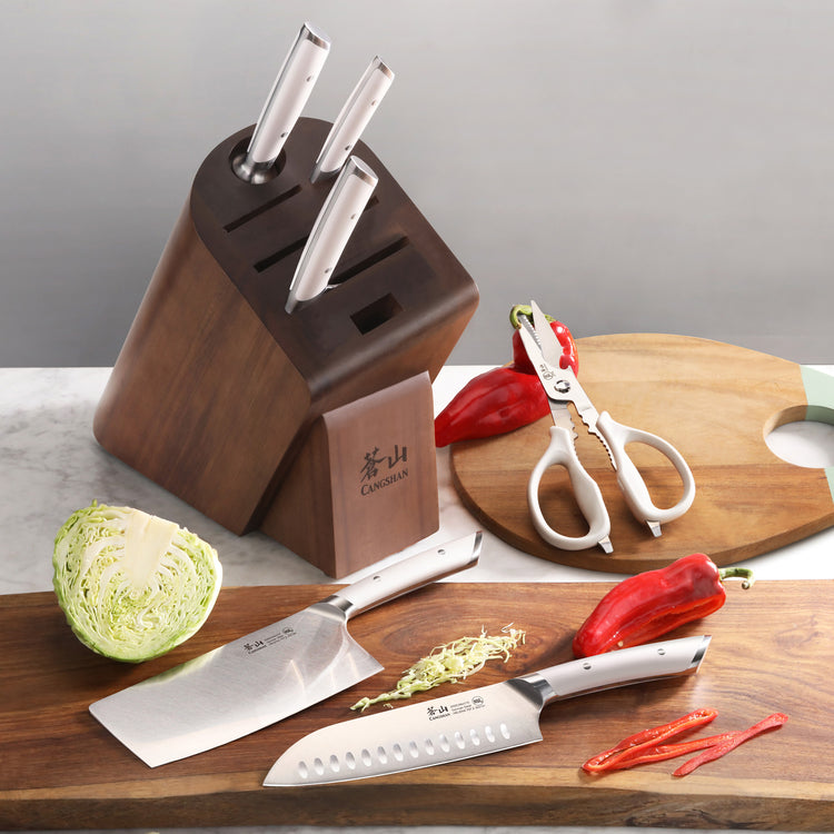 Wooden knife block with three knives inserted, two knives and a pair of kitchen scissors on a cutting board, surrounded by sl