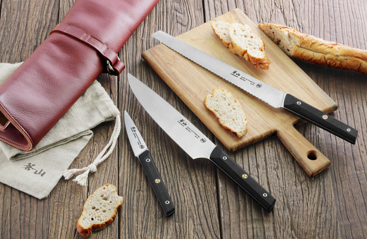 Four kitchen knives with black handles displayed on a wooden surface, alongside sliced bread, a cutting board, and a rolled l