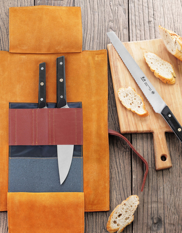 Two kitchen knives with black handles in a tan leather roll next to a serrated knife and sliced bread on a wooden cutting boa