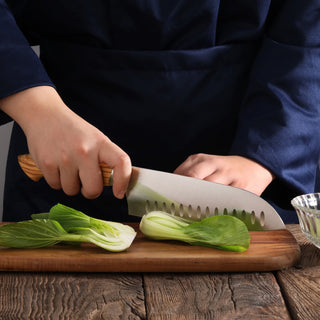 
                    
                      Person slicing bok choy on wooden board with a 7-inch Santoku knife featuring a wooden handle and scalloped blade.
                    
                  