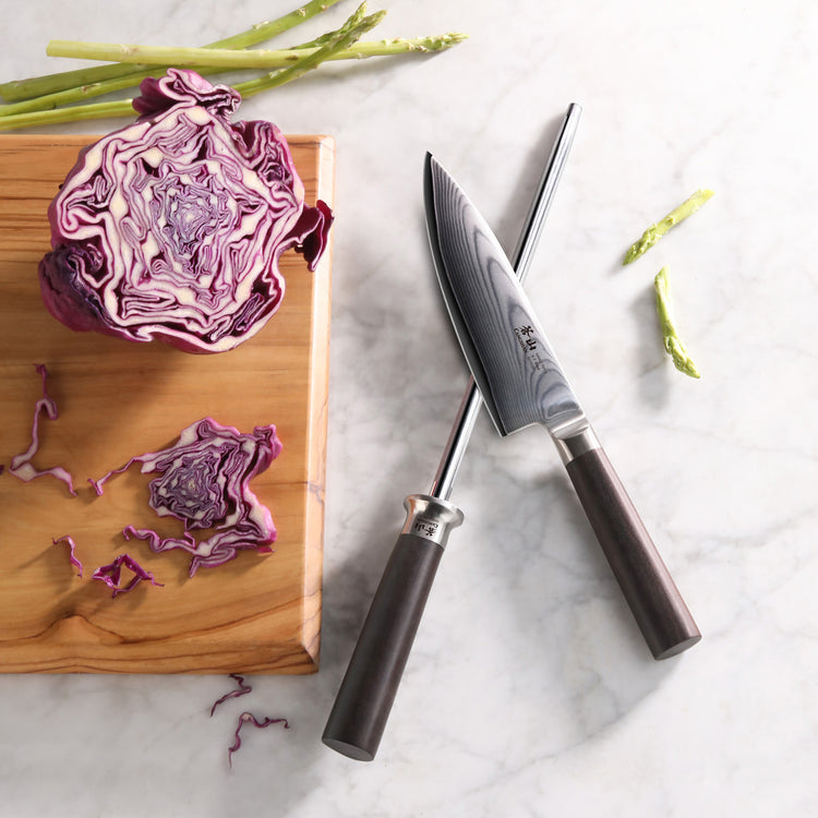 6-inch chef's knife and sharpening steel crossed on marble surface next to wooden board with halved red cabbage and asparagus