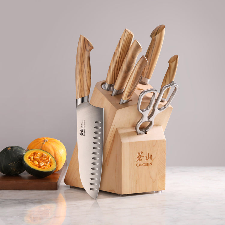 Knife block set with six knives and kitchen scissors, wooden handles and block, on marble surface with cut pumpkins nearby.
