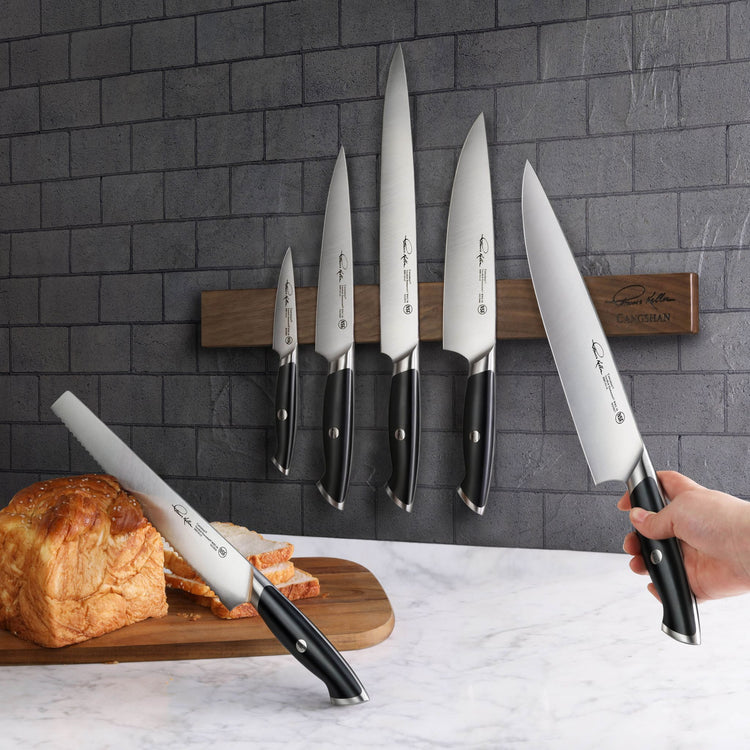 Seven black-handled knives with stainless steel blades, five hanging on a wooden magnetic strip and two held near a loaf of b