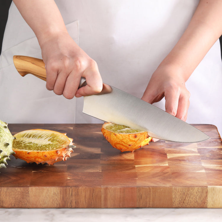 Hand holding a knife with a maple handle, slicing a horned melon on a wooden cutting board.