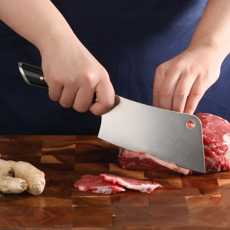 Hands slicing raw meat with a cleaver knife on a wooden cutting board with ginger root nearby.