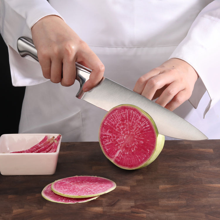 Person slicing a round pink radish with a large chef's knife on a wooden cutting board.