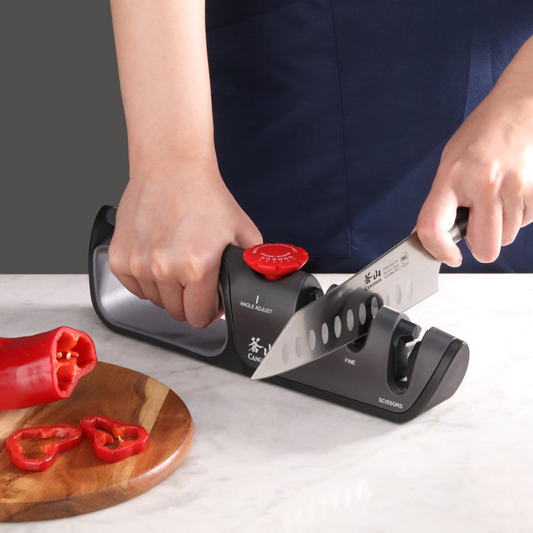 Hands using a three-slot knife and scissor sharpener with a red adjustment knob on a countertop.