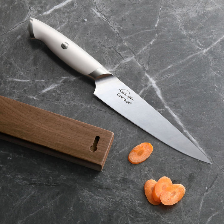 White-handled kitchen knife with stainless steel blade next to wooden magnetic knife bar and sliced carrots on gray marble su