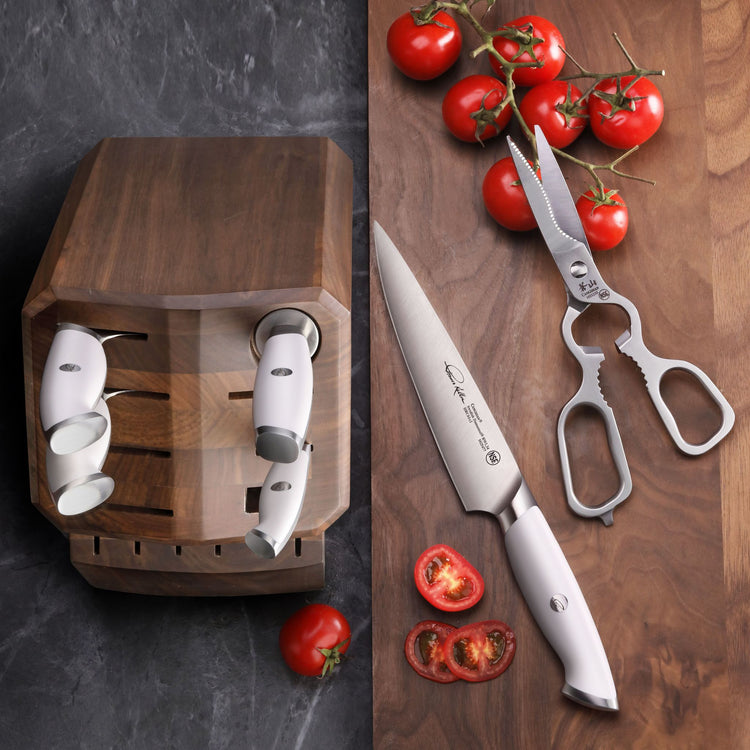 Wooden knife block with four knives inserted, a chef's knife and kitchen shears on a wooden surface with fresh tomatoes.